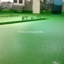 Świeżo natryśnięta, zielona izolacja pianką poliuretanową na podłodze hali, widoczne dysze natryskowe i logo firmy izolacyjnej w tle.