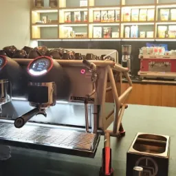 Profesjonalny ekspres do kawy Sanremo z dwoma grupami i wyświetlaczami, obok młynek do kawy, w tle półki z zapasami kawy i inny ekspres.