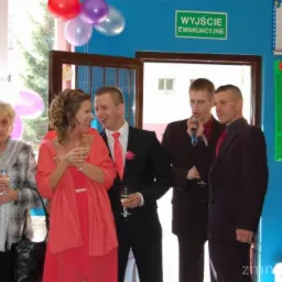 Zespół muzyczny na imprezie, wokalista śpiewa do mikrofonu. W tle dekoracje z balonów, na ścianie tablica z cennikiem. Impreza okolicznościowa w lokalu.