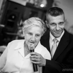 Czarno-białe zdjęcie kobiety śpiewającej do mikrofonu z uśmiechniętym mężczyzną w garniturze w tle, prawdopodobnie podczas uroczystości.