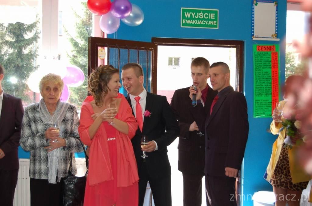 Zespół muzyczny na imprezie, wokalista śpiewa do mikrofonu. W tle dekoracje z balonów, na ścianie tablica z cennikiem. Impreza okolicznościowa w lokalu.