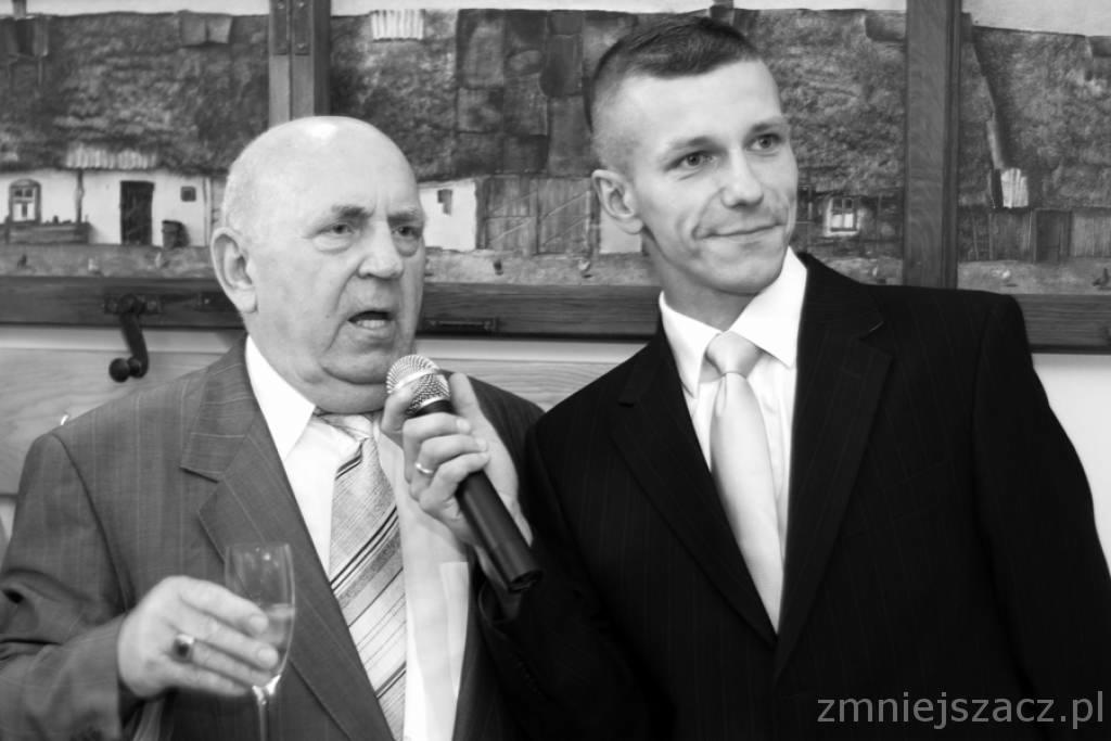 Czarno-białe zdjęcie dwóch mężczyzn w garniturach. Jeden trzyma mikrofon i szklankę, drugi stoi obok. W tle obraz z motywem wiejskim.