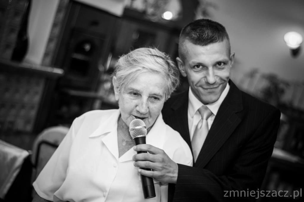 Czarno-białe zdjęcie kobiety śpiewającej do mikrofonu z uśmiechniętym mężczyzną w garniturze w tle, prawdopodobnie podczas uroczystości.