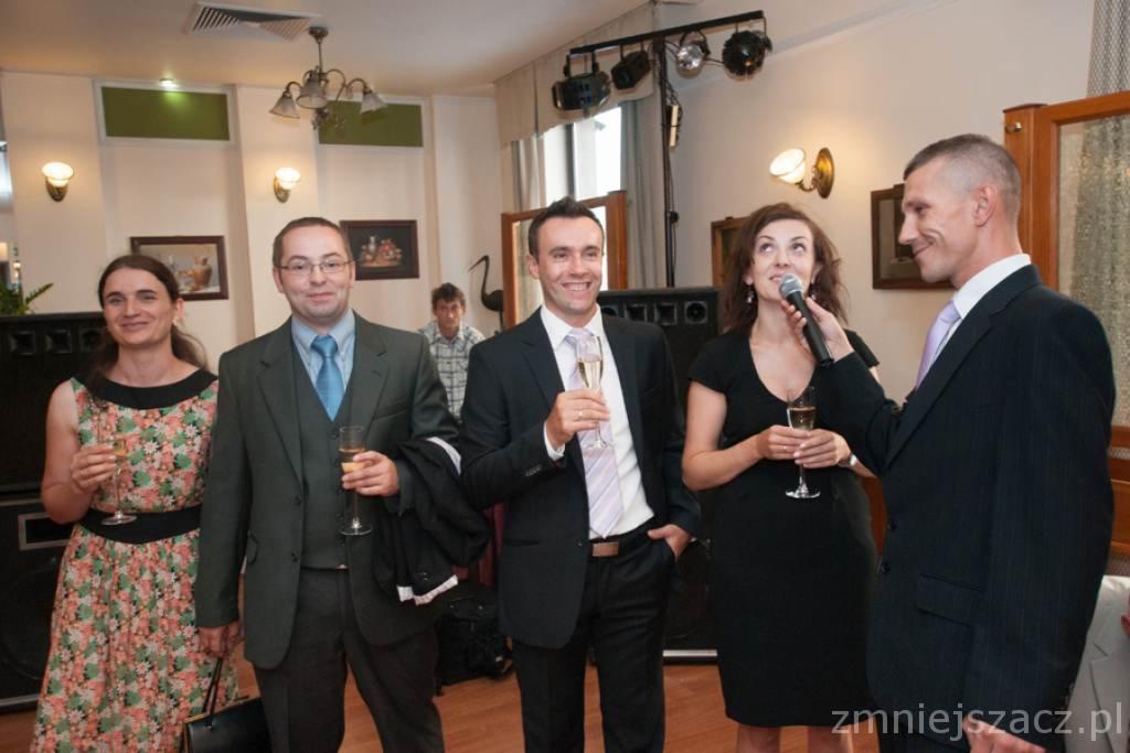 Konferansjer z mikrofonem wita gości weselnych, wznosząc toast szampanem. W tle widoczny sprzęt nagłośnieniowy i oświetlenie.