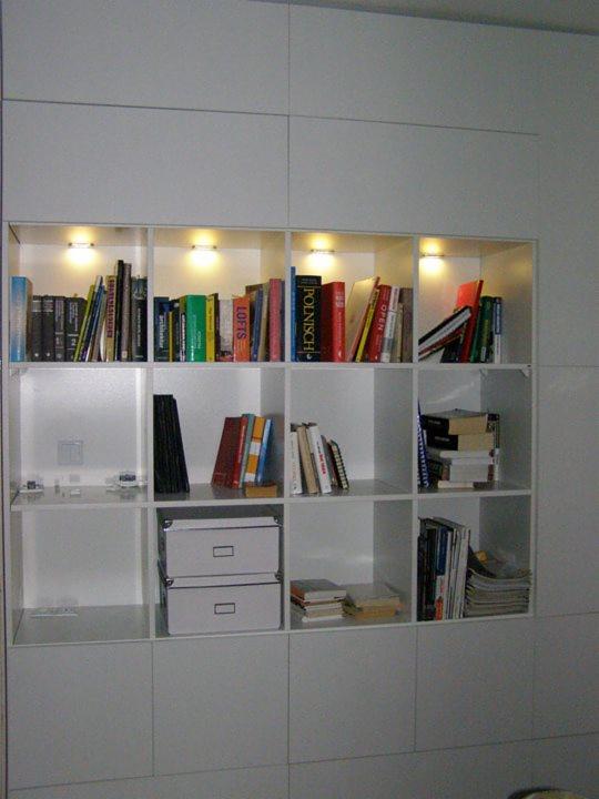 Biała zabudowa meblowa z półkami, podświetlane wnęki z książkami i dekoracjami, dolne szafki z metalowymi pudełkami, minimalistyczny design.