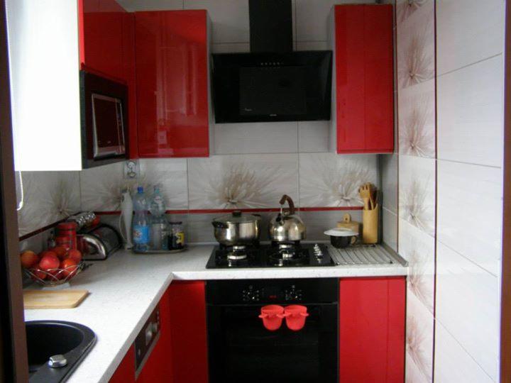 Kompaktowa kuchnia z czerwonymi szafkami na wysoki połysk i białym blatem, czarna płyta grzewcza i piekarnik, wzorzyste płytki ścienne, naczynia i akcesoria na blacie.