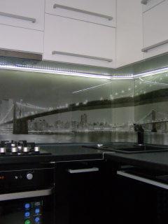 Nowoczesna kuchnia z czarno-białym panelem szklanym nad blatem przedstawiającym most Brookliński, białe szafki górne z podświetleniem LED, czarne szafki dolne i sprzęt AGD.