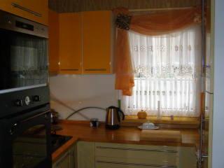 Żółte szafki kuchenne z połyskiem, okno z firanką i pomarańczową dekoracją, czajnik elektryczny na blacie, częściowo widoczny piekarnik.