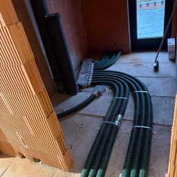 MAX PROJEKT SPÓŁKA Z OGRANICZONĄ ODPOWIEDZIALNOŚCIĄ - Instalacja rekuperacji podłogowej w surowym wnętrzu domu w Henrykowie-Uroczem. Widoczne rury wentylacyjne i rozdzielacz. Ściany z cegły dziurawki.