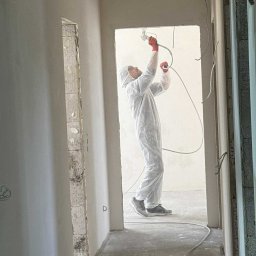 MAX PROJEKT SPÓŁKA Z OGRANICZONĄ ODPOWIEDZIALNOŚCIĄ - Pracownik w kombinezonie ochronnym maluje ściany natryskowo wewnątrz remontowanego pomieszczenia. Widoczne surowe ściany i przewody.