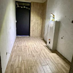 MAX PROJEKT PIOTR DMOWSKI - Wykończona łazienka z drewnopodobną podłogą i częściowo wykończonymi ścianami. Widoczna czarna rama drzwi i zabudowa pod umywalkę z narzędziami.