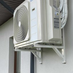 MAX PROJEKT SPÓŁKA Z OGRANICZONĄ ODPOWIEDZIALNOŚCIĄ - Biały klimatyzator Kaisai zamontowany na ścianie budynku, widoczne rury w izolacji. Montaż na metalowym wsporniku, okno w tle. Zewnętrzna jednostka klimatyzacji.