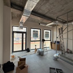 Wnętrze w trakcie montażu instalacji wentylacyjnej z widocznymi kanałami, drabina i surowy betonowy strop. Widoczne okna z widokiem na zewnątrz.