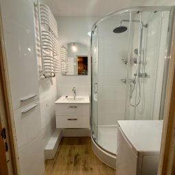 Biała łazienka z kabiną prysznicową, umywalką z szafką i grzejnikiem drabinkowym. Uwagę zwraca drewnopodobna podłoga i odbicie osoby w lustrze.
