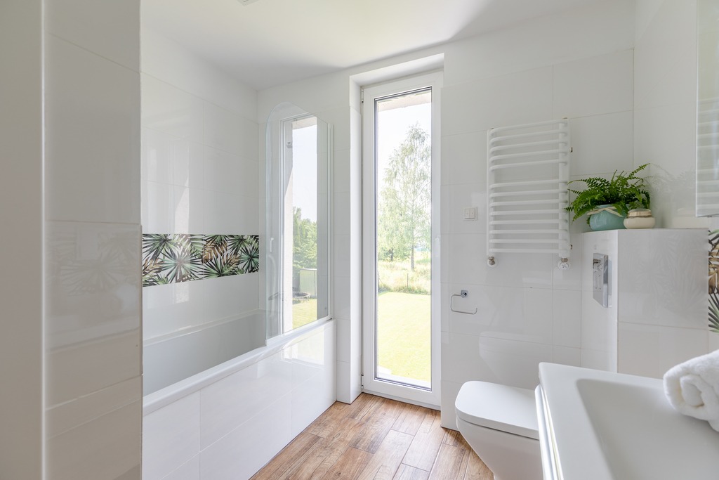 Jasna łazienka z wanną, kabiną prysznicową i oknem z widokiem na zieleń. Białe płytki z motywem roślinnym, drewnopodobna podłoga, grzejnik drabinkowy.