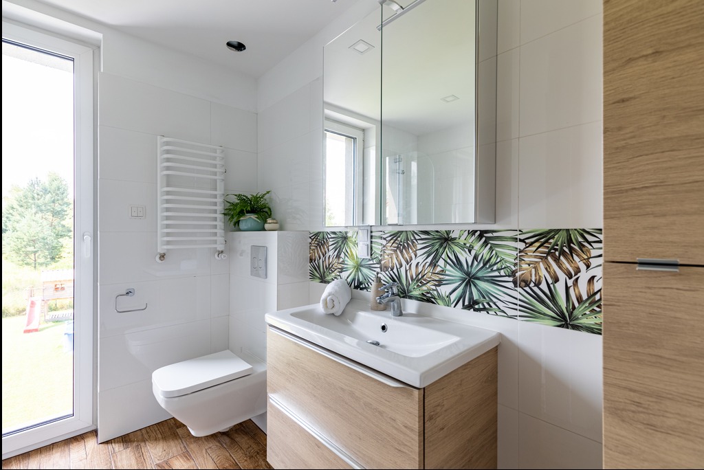 Jasna łazienka z białymi płytkami, drewnianą szafką pod umywalką i akcentem tropikalnych liści na ścianie. Widoczne okno i grzejnik drabinkowy.