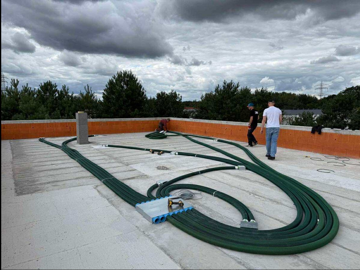 Montaż instalacji rekuperacji na dachu budynku w trakcie budowy. Widoczne zielone rury wentylacyjne, pracownicy i fragment muru. Pochmurne niebo w tle.