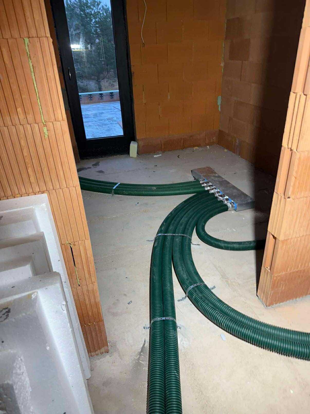 Zielone rury rekuperacji ułożone na podłodze w surowym wnętrzu z ceglanymi ścianami i oknem balkonowym. Widoczny etap instalacji systemu wentylacji.