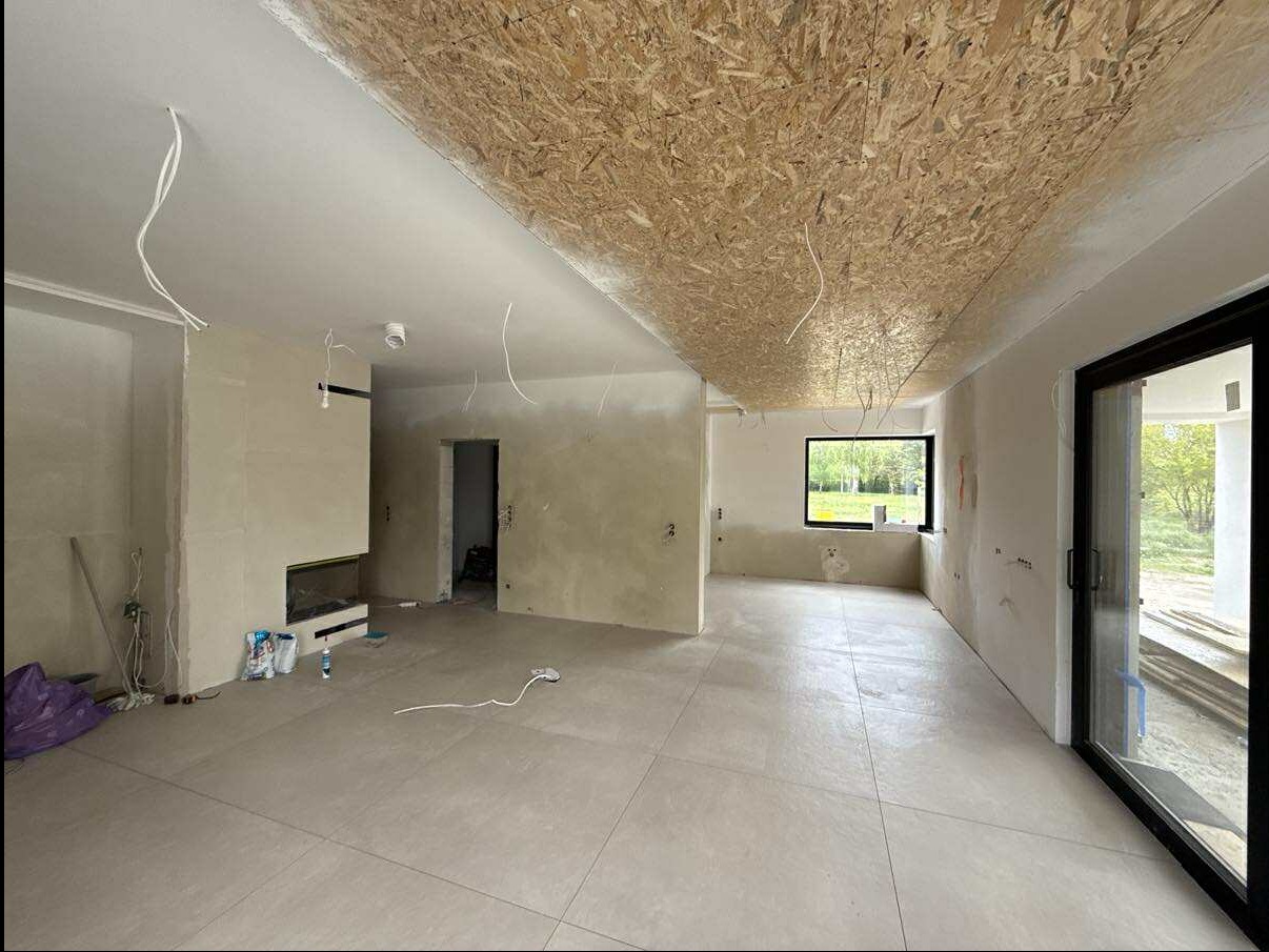 Wnętrze w trakcie prac wykończeniowych: surowy strop OSB, gładzie na ścianach, ułożona podłoga z płytek, okno z widokiem na zieleń, przygotowane miejsce na kominek.