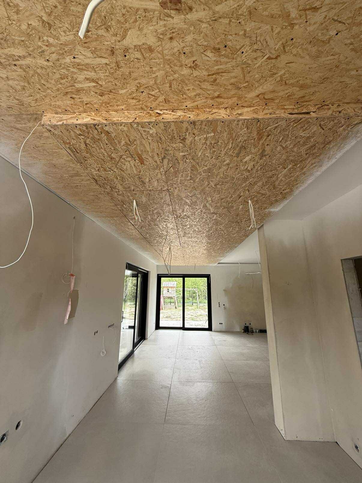 Wnętrze w trakcie wykończenia: sufit z płyty OSB, widoczne przewody, ściany przygotowane do malowania, szare płytki podłogowe, okno balkonowe z widokiem na zieleń.