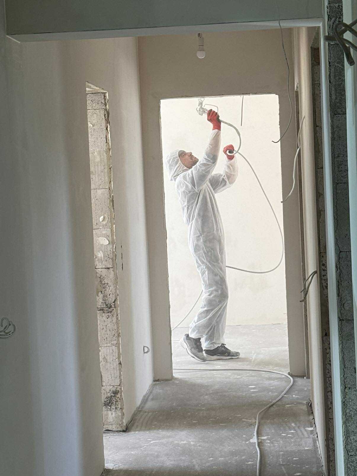 Pracownik w kombinezonie ochronnym maluje ściany natryskowo wewnątrz remontowanego pomieszczenia. Widoczne surowe ściany i przewody.