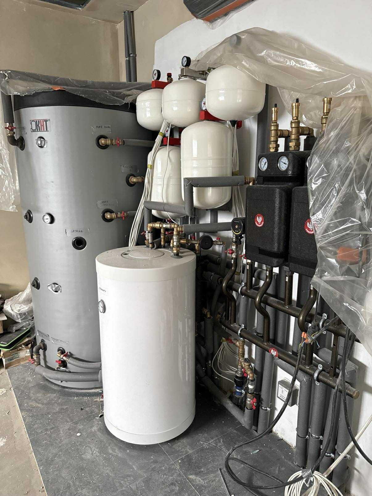 Kompleksowa instalacja grzewcza z zbiornikami, pompami i rurami w pomieszczeniu technicznym. Widoczne elementy izolacji i manometry.