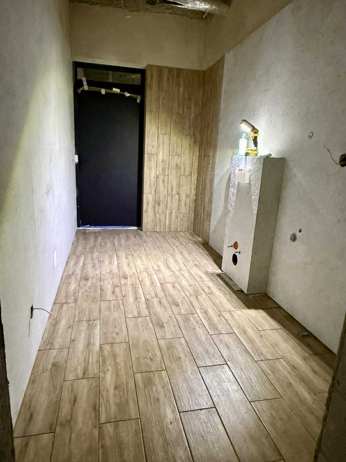 Wykończona łazienka z drewnopodobną podłogą i częściowo wykończonymi ścianami. Widoczna czarna rama drzwi i zabudowa pod umywalkę z narzędziami.