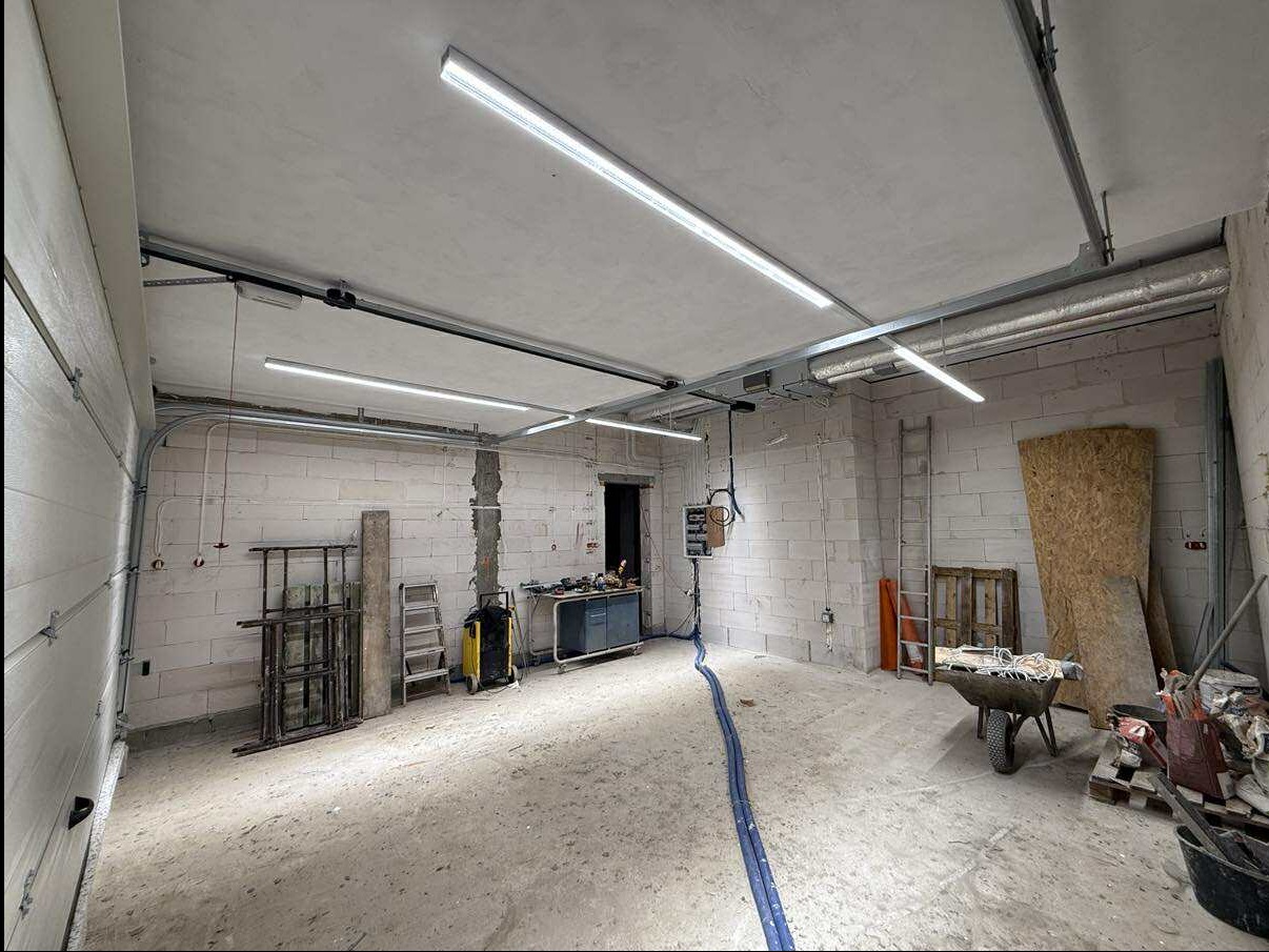 Wnętrze garażu w trakcie remontu: surowe ściany z betonu komórkowego, nowe oświetlenie LED, narzędzia i materiały budowlane. Widoczny wózek i drabina.