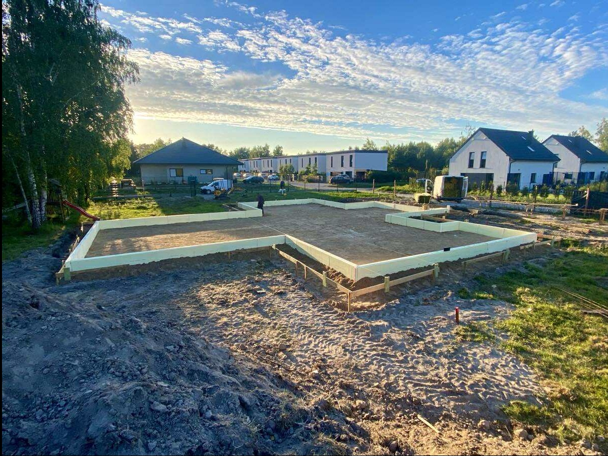 Fundamenty domu jednorodzinnego w trakcie budowy, ocieplone styropianem, widok z góry na plac budowy w słoneczny dzień. W tle zabudowa jednorodzinna i drzewa.