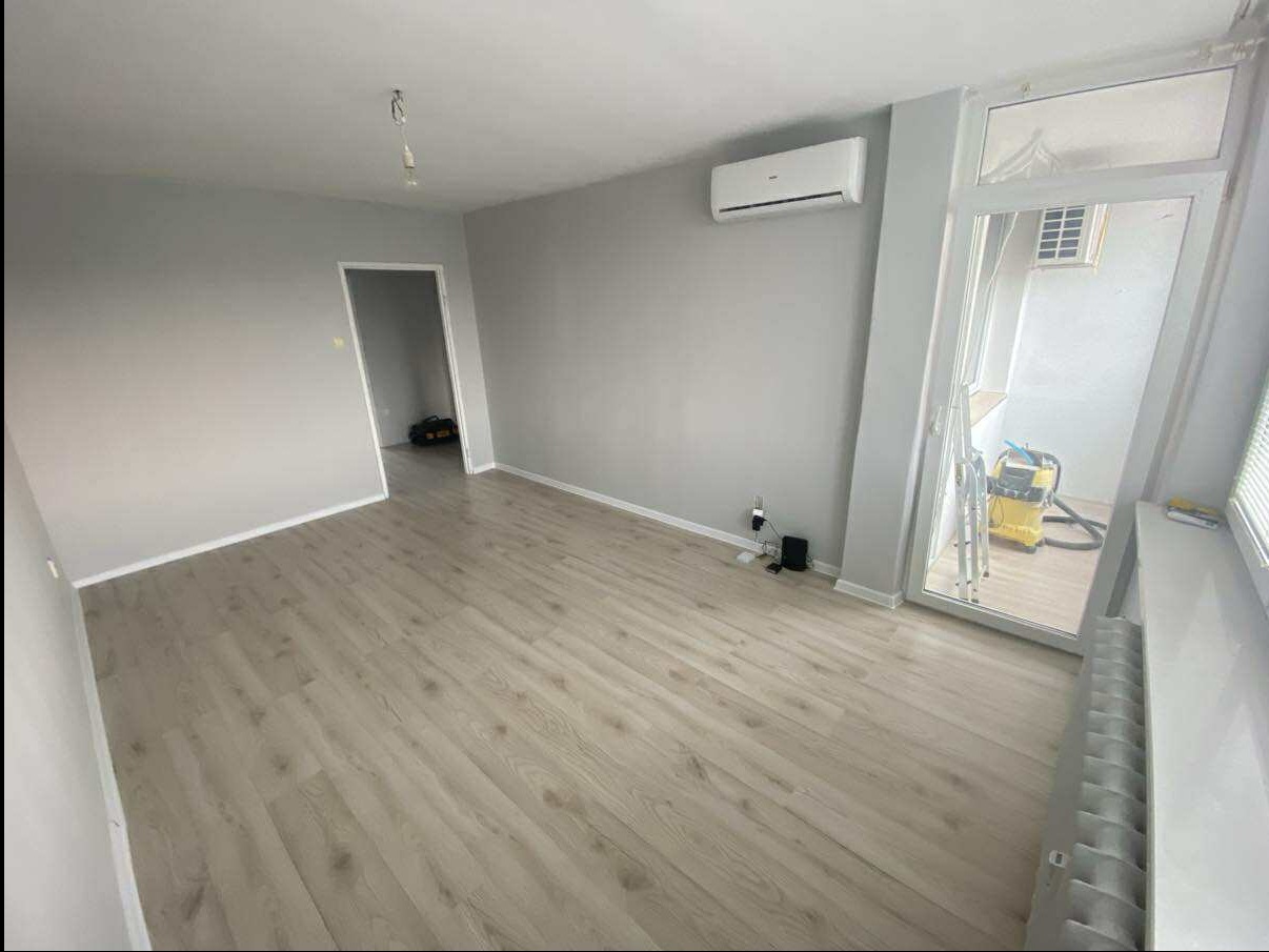 Jasne, szare wnętrze po remoncie: panele podłogowe, świeżo pomalowane ściany, klimatyzacja, okno balkonowe z widokiem na balkon. Widoczny odkurzacz przemysłowy.