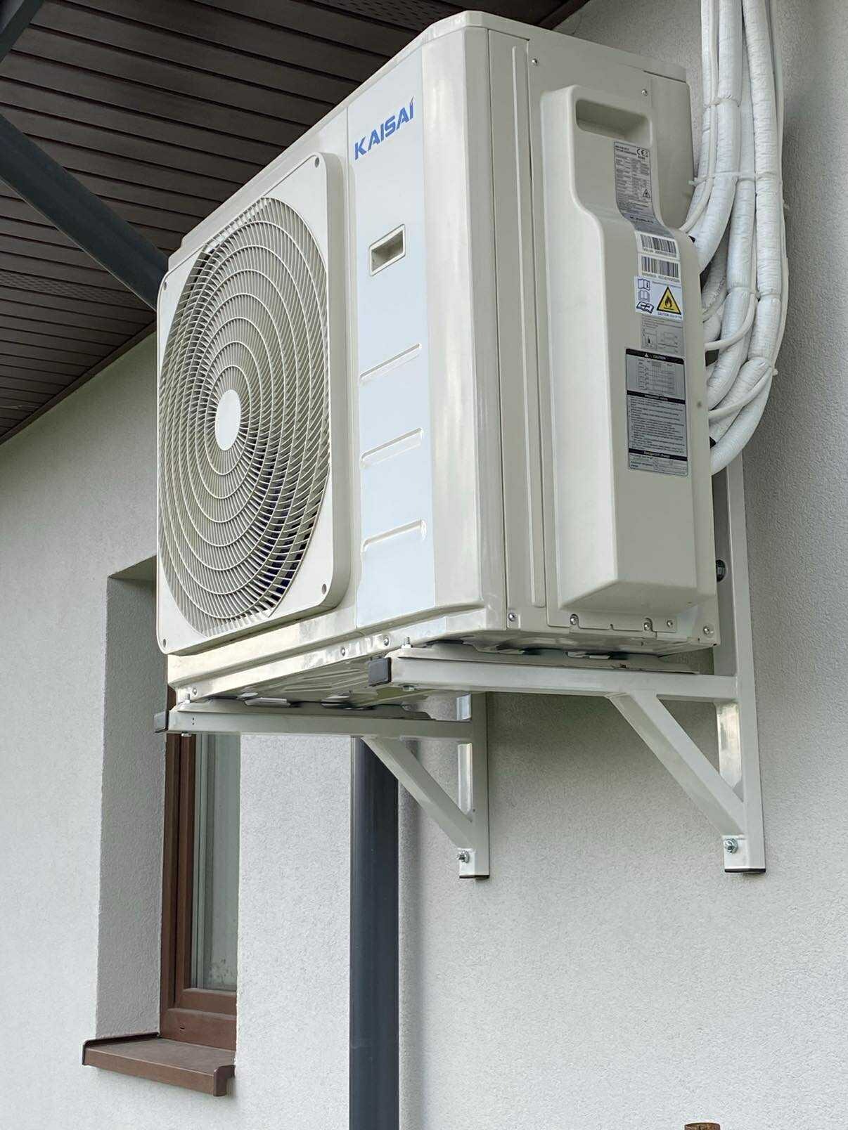 Biały klimatyzator Kaisai zamontowany na ścianie budynku, widoczne rury w izolacji. Montaż na metalowym wsporniku, okno w tle. Zewnętrzna jednostka klimatyzacji.