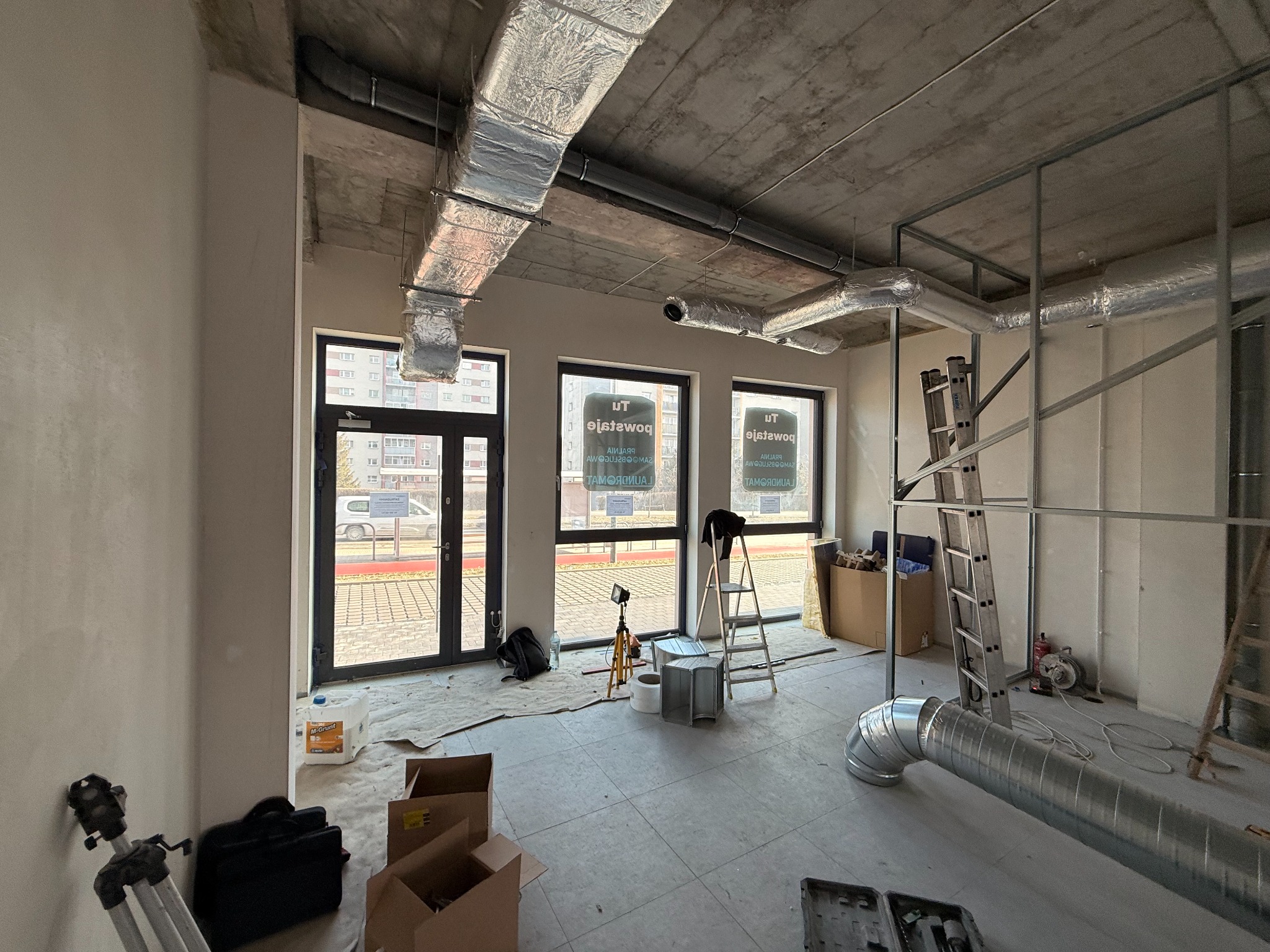 Wnętrze w trakcie montażu instalacji wentylacyjnej z widocznymi kanałami, drabina i surowy betonowy strop. Widoczne okna z widokiem na zewnątrz.