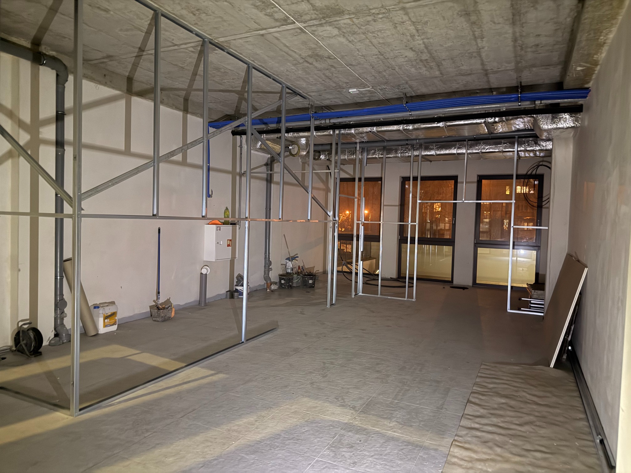 Wnętrze w trakcie prac wykończeniowych: metalowa konstrukcja ścian działowych, instalacje pod sufitem, surowy betonowy strop, płytki na podłodze. Widoczne narzędzia i materiały budowlane.
