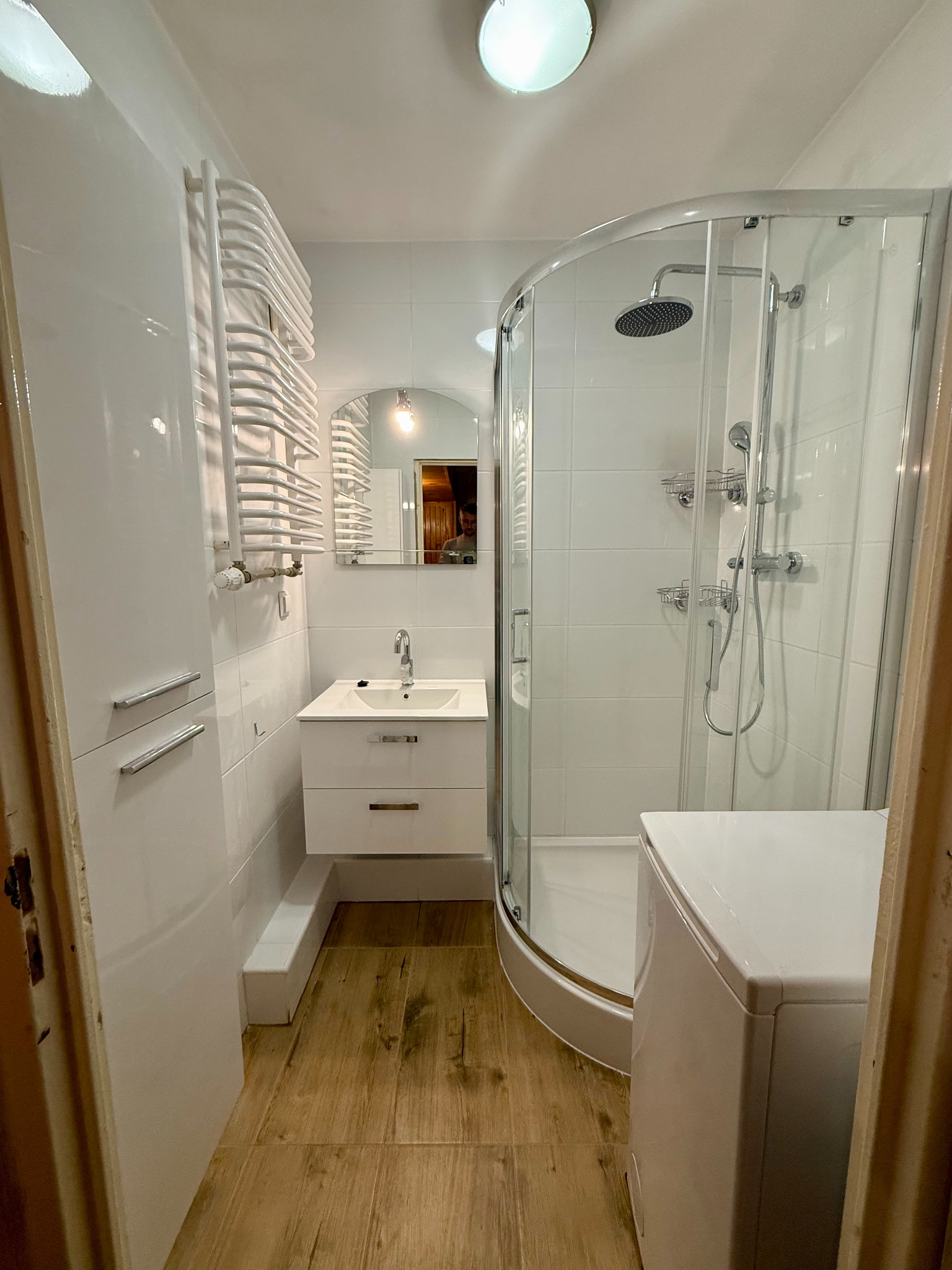 Biała łazienka z kabiną prysznicową, umywalką z szafką i grzejnikiem drabinkowym. Uwagę zwraca drewnopodobna podłoga i odbicie osoby w lustrze.