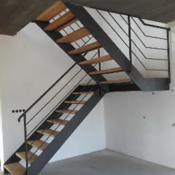 Metalowe schody z drewnianymi stopniami w surowym wnętrzu. Ciemna konstrukcja kontrastuje z jasnymi ścianami i schodami. Widoczne otwory na gniazdka i niedokończone wykończenie.