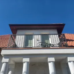 Balkon z szarą balustradą i kutymi elementami dekoracyjnymi w kształcie liści na tle błękitnego nieba i czerwonej dachówki.