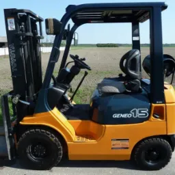 Żółty wózek widłowy Toyota Geneo 15 z czarną kabiną, widoczny z boku na szarej nawierzchni, z polem uprawnym w tle.