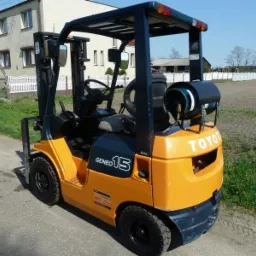 Pomarańczowo-granatowy wózek widłowy Toyota Geneo 15 z butlą gazową z tyłu, stojący na asfaltowej drodze z budynkiem i polem w tle.