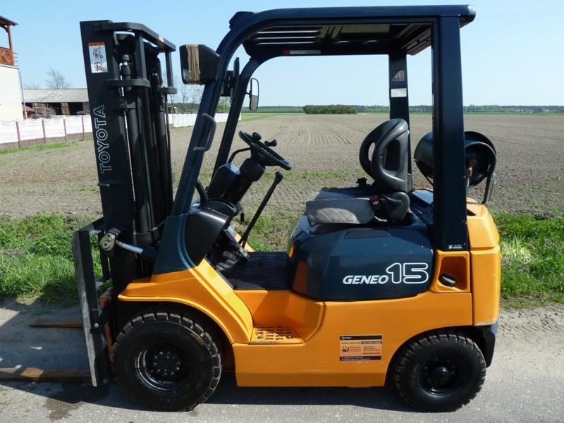 Żółty wózek widłowy Toyota Geneo 15 z czarną kabiną, widoczny z boku na szarej nawierzchni, z polem uprawnym w tle.