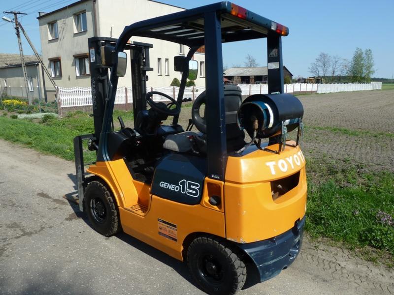 Pomarańczowo-granatowy wózek widłowy Toyota Geneo 15 z butlą gazową z tyłu, stojący na asfaltowej drodze z budynkiem i polem w tle.