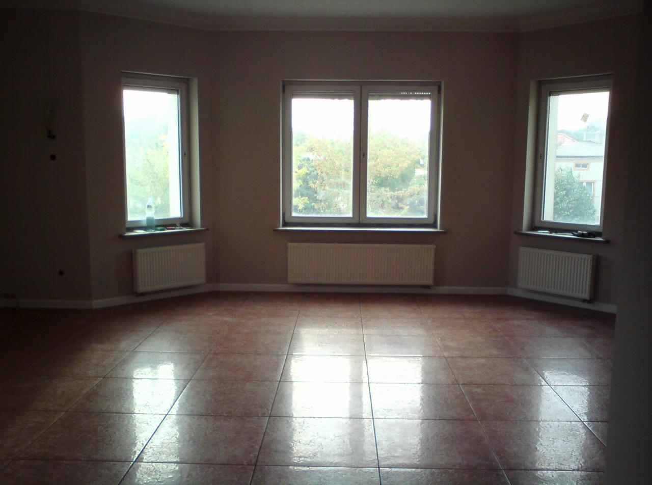 Puste, jasne pomieszczenie z brązowymi płytkami podłogowymi, trzema oknami z widokiem na zieleń i grzejnikami pod każdym z nich. Ściany w kolorze beżowym.