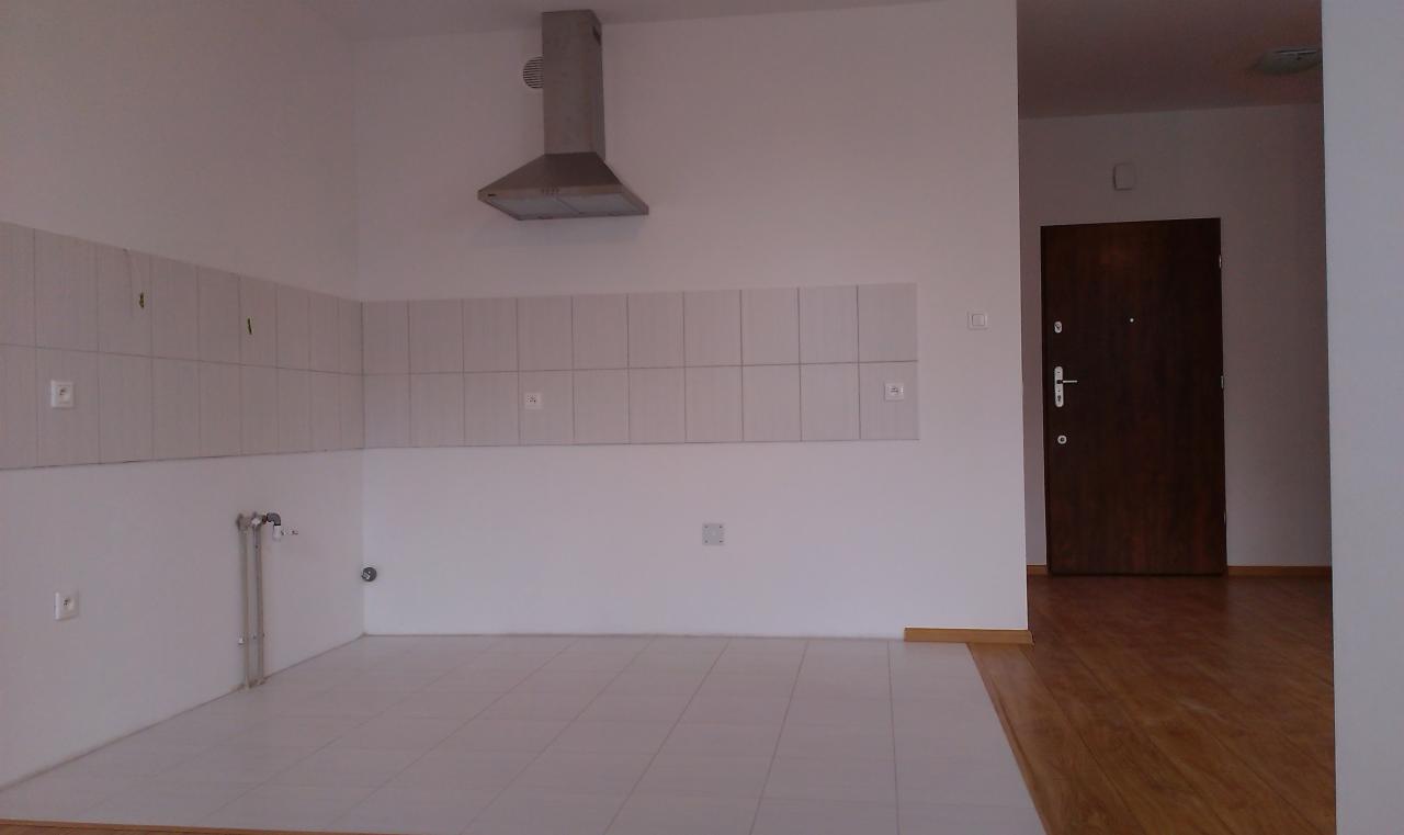 Pusta kuchnia po remoncie: białe płytki na ścianach i podłodze, zamontowany okap, widoczne przyłącza wodne, brązowe drzwi wejściowe.