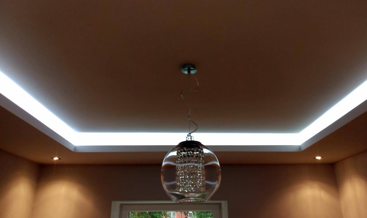 Podwieszany sufit z oświetleniem LED wzdłuż krawędzi, uzupełniony wiszącą, okrągłą lampą z kryształkami, w tle ściana w odcieniach brązu.