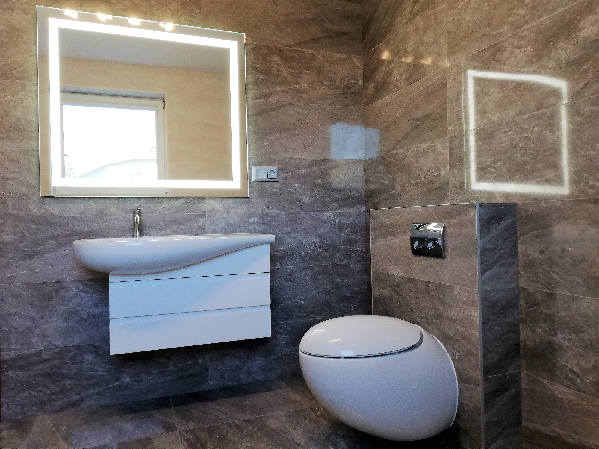 Nowoczesna łazienka z marmurowymi płytkami, podświetlanym lustrem, wiszącą umywalką z szafką i toaletą w minimalistycznym stylu.