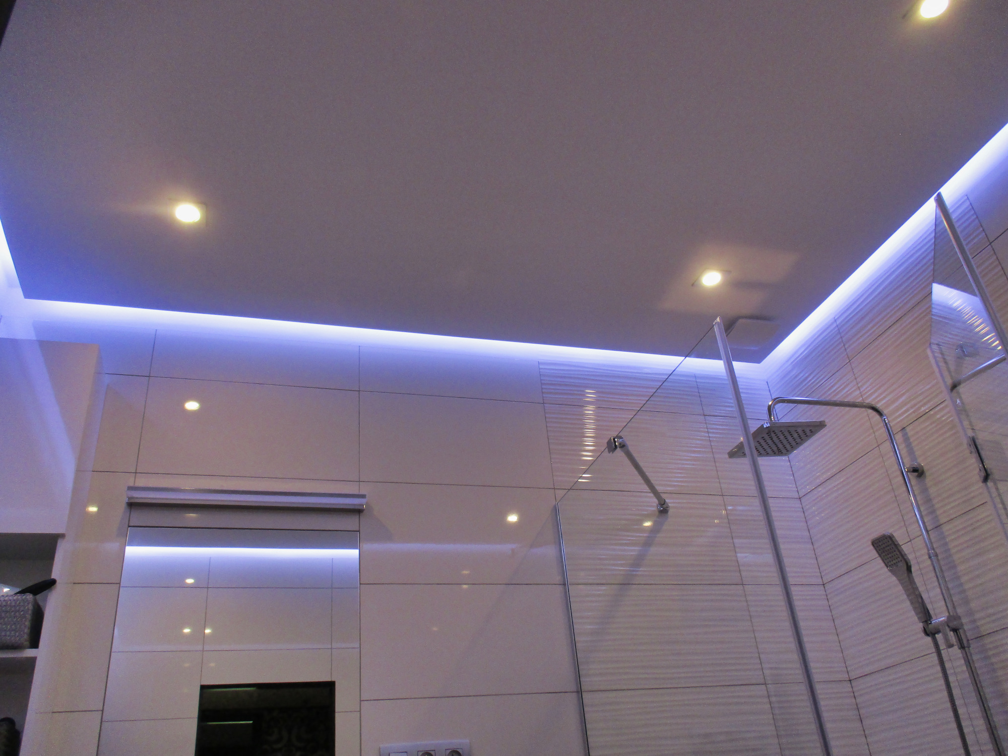 Nowoczesna kabina prysznicowa z deszczownicą i oświetleniem LED, wykończona białymi płytkami z delikatnym wzorem, widziana z perspektywy dolnej.