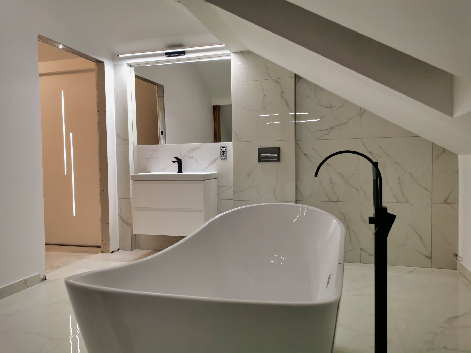 Nowoczesna łazienka w odcieniach bieli z wanną wolnostojącą i armaturą w kolorze czarnym, marmurowe płytki na ścianach i podłodze, widoczny fragment oświetlonej wnęki drzwiowej.