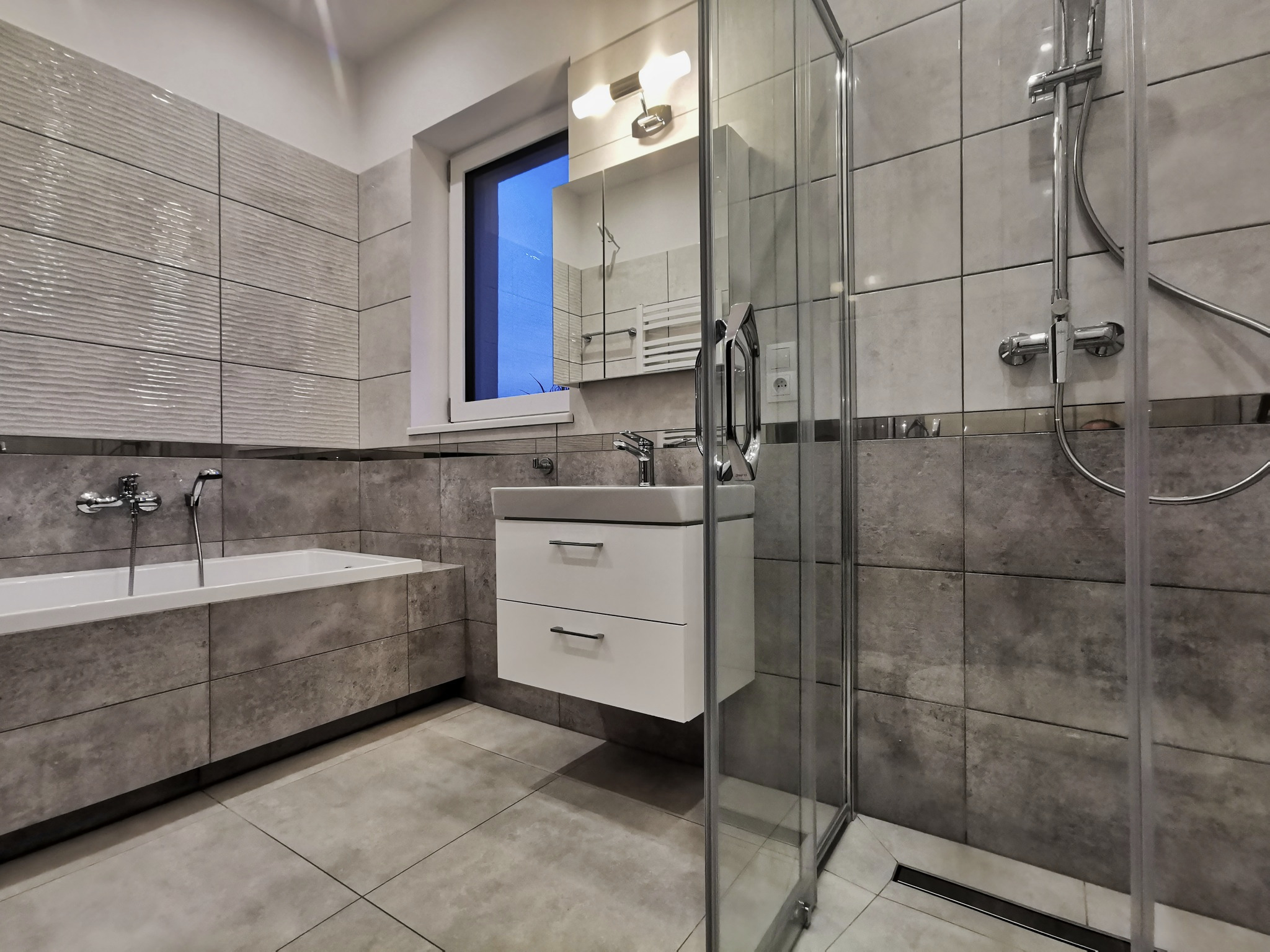 Nowoczesna łazienka w Łodzi z kabiną prysznicową typu walk-in, umywalką z szafką podwieszaną i wanną obudowaną płytkami imitującymi beton, oświetlona światłem lampy nad lustrem.