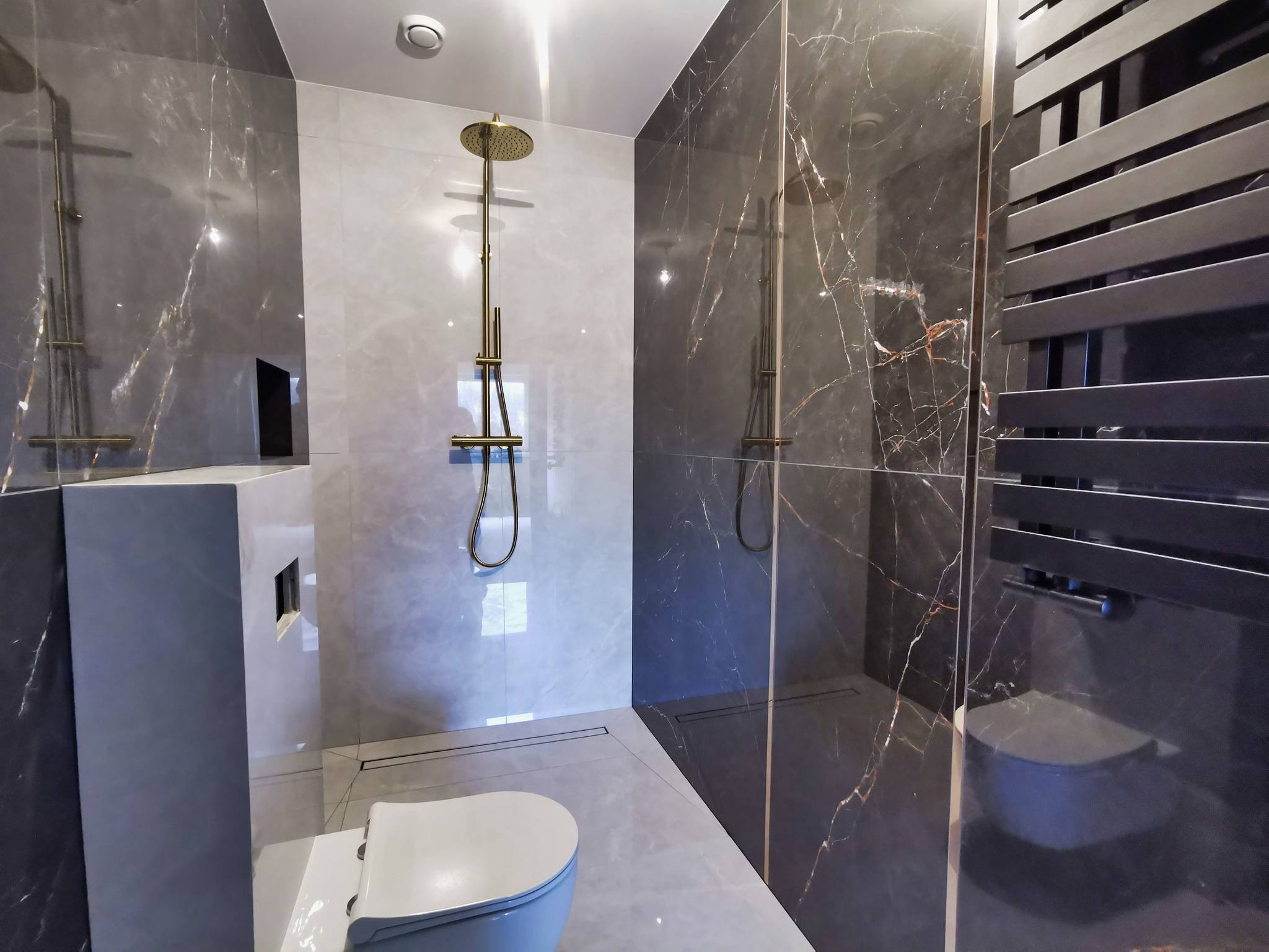 Nowoczesna łazienka z szarymi marmurkowymi płytkami, złotą deszczownicą i czarnym grzejnikiem drabinkowym, widoczny odpływ liniowy w podłodze.