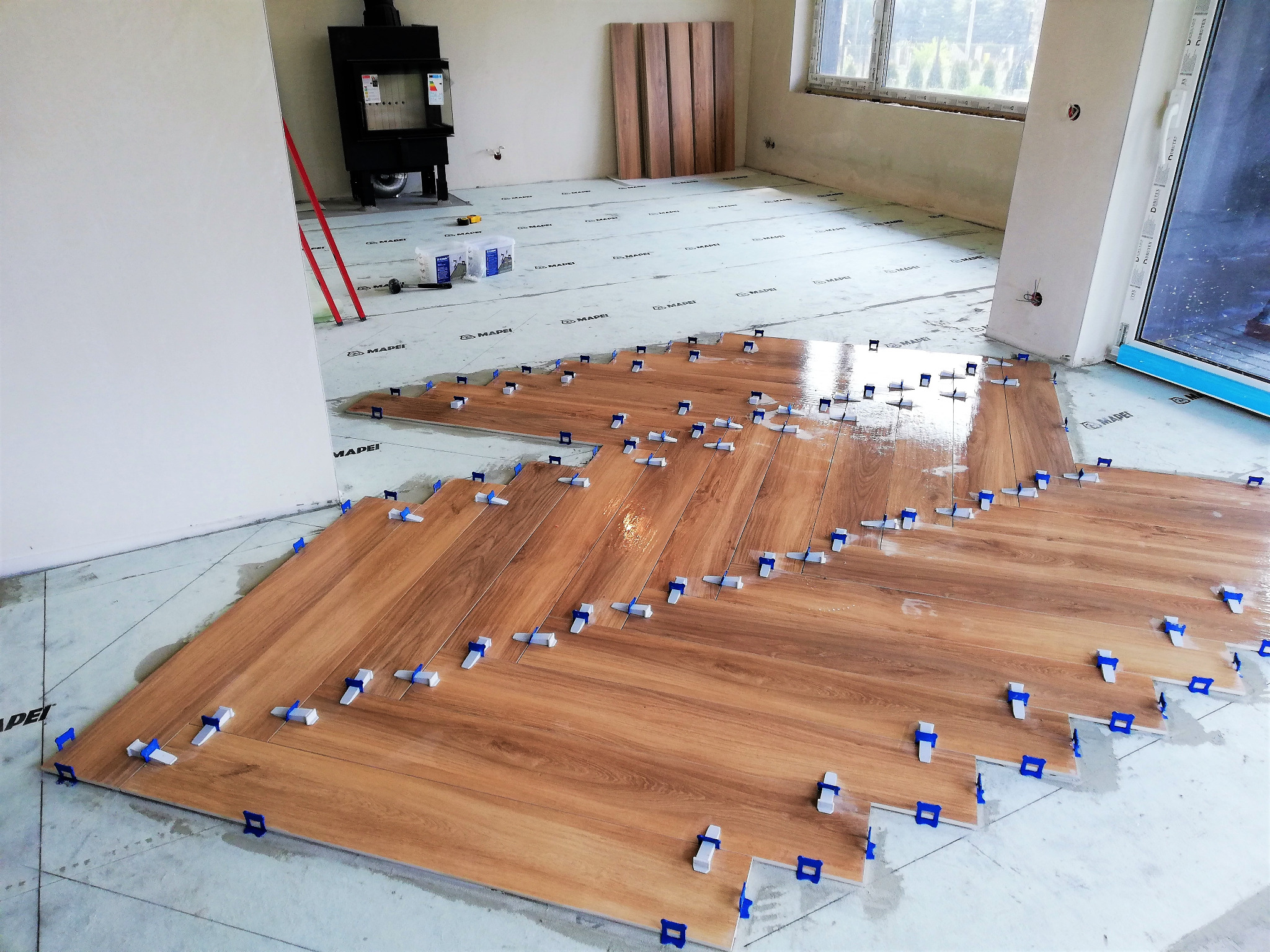 Układanie drewnopodobnych płytek podłogowych w jodełkę, z użyciem niebieskich krzyżyków dystansowych, na podkładzie z folii. W tle widoczne materiały budowlane i piec.