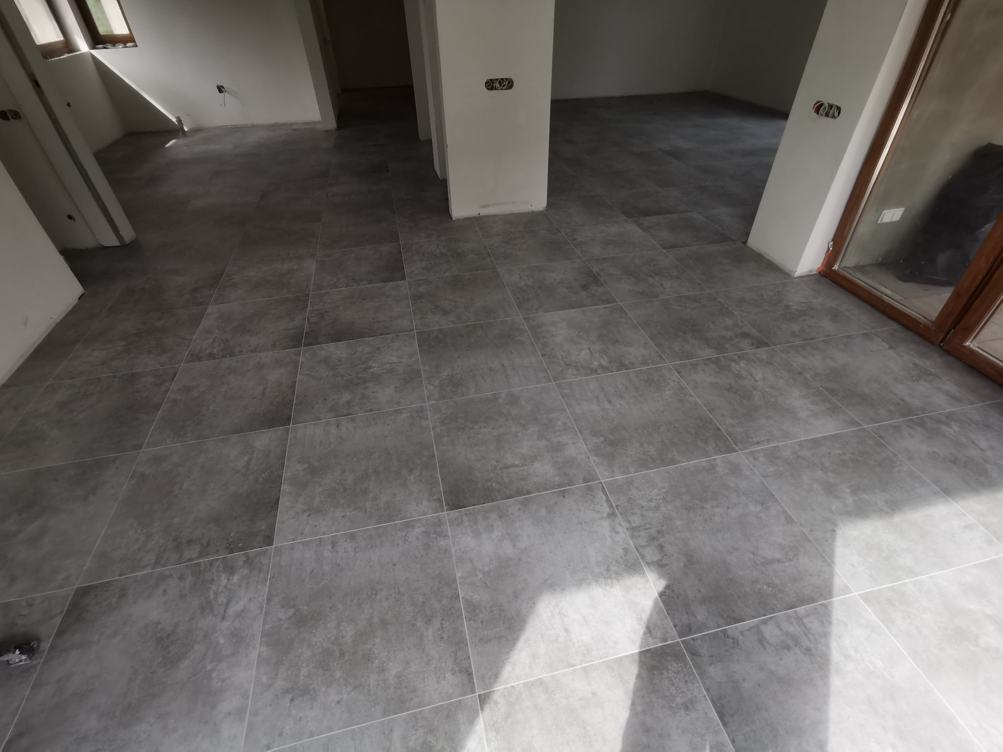 Ułożone, szare płytki podłogowe w przestronnym, jasnym pomieszczeniu z białymi ścianami i widocznymi instalacjami elektrycznymi.